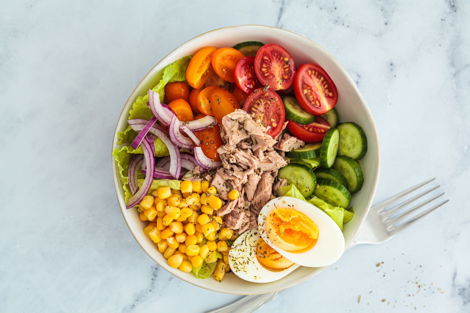 A salad with tuna, eggs, tomatoes, lettuce, cucumbers, corn, and onion.