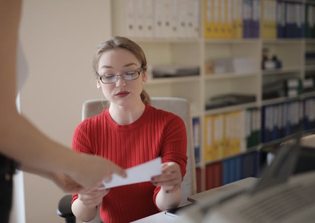 A woman wearing glasses is holding a piece of paper, looking focused and engaged in what she's reading.