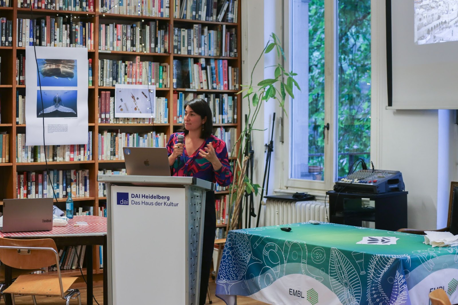 A woman scientist stands at a podium inside a library, delivering a talk. The podium has the logo of DAI Heidelberg on it.