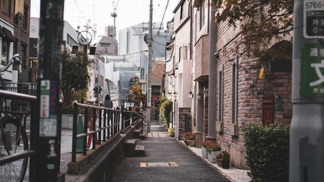 Narrow city street with a sidewalk lined by buildings, including brick facades crafted by skilled architects. Distant modern skyscrapers and construction cranes hint at the ongoing community development.