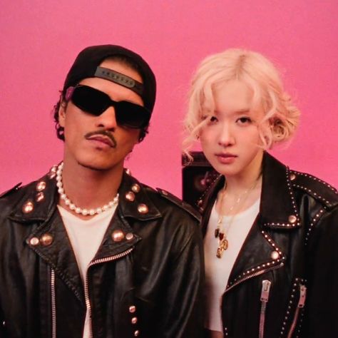 This contains an image of Bruno Mars and Rosé standing next to each other in front of a pink background wearing a black leather jackets