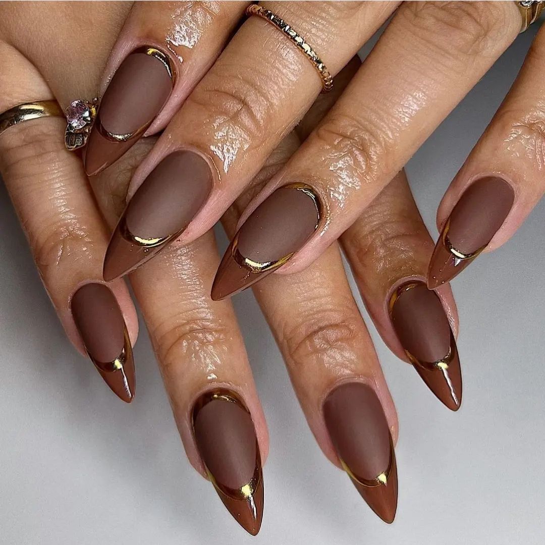 Glossy taupe stiletto nails with gold accents at the base, displayed on a hand against a neutral background.

