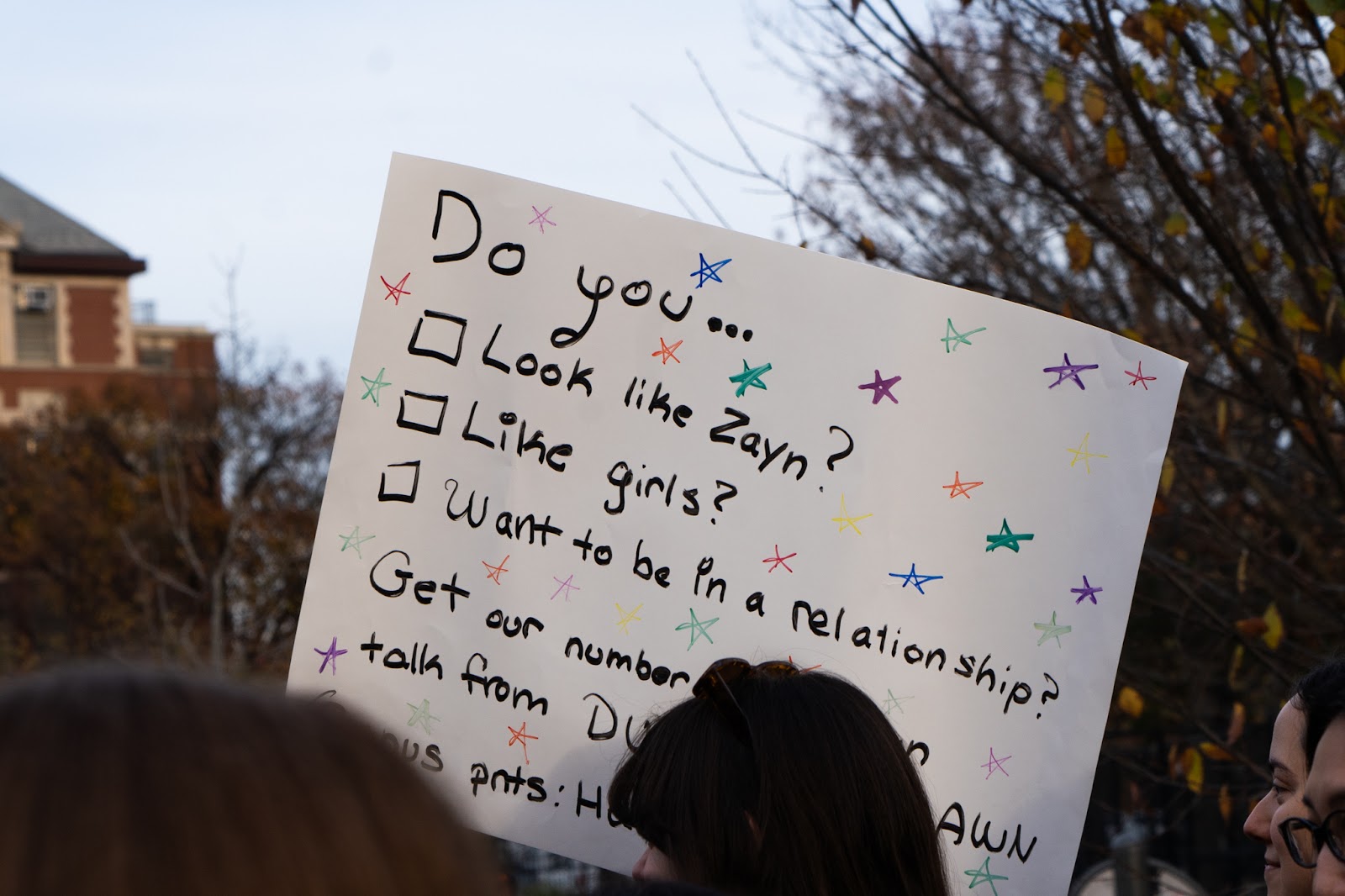 An image of a crowd member from the  Zayn Malik look-alike contest holding up a sign that says “Do you look like Zayn? Like girls? Want to be in a relationship? Get our number so we can talk from Dusk till Dawn”.
