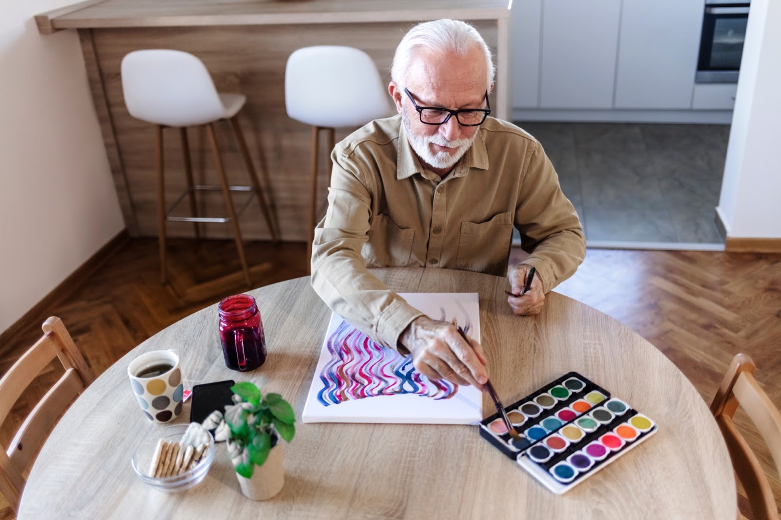 An older adult man painting with watercolors at his kitchen table.