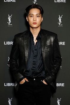 This contain EXO Kai' in a black suit and tie standing next to a sign that says libre