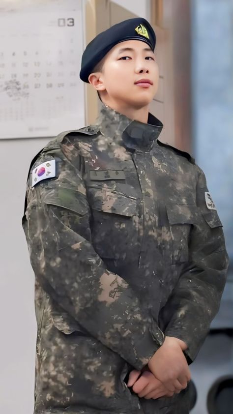 This contain BTS RM   in uniform standing next to a wall