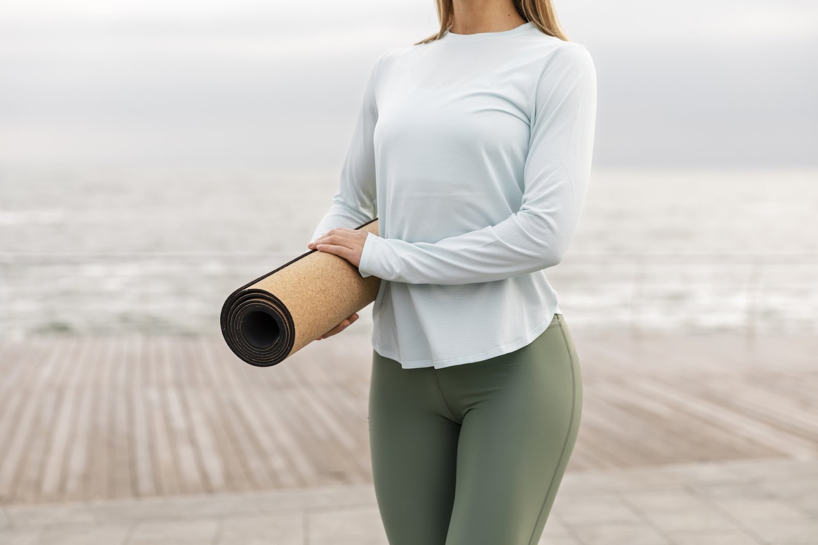 who invented yoga pants
An individual is standing outdoors near a body of water, holding a rolled-up yoga mat. They are wearing a light blue long-sleeve shirt and green leggings. The background features a wooden boardwalk and a cloudy sky.