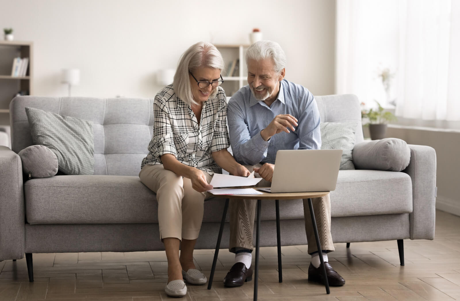 An older couple working together on the couch and smiling while looking through paperwork to learn the costs of senior living.