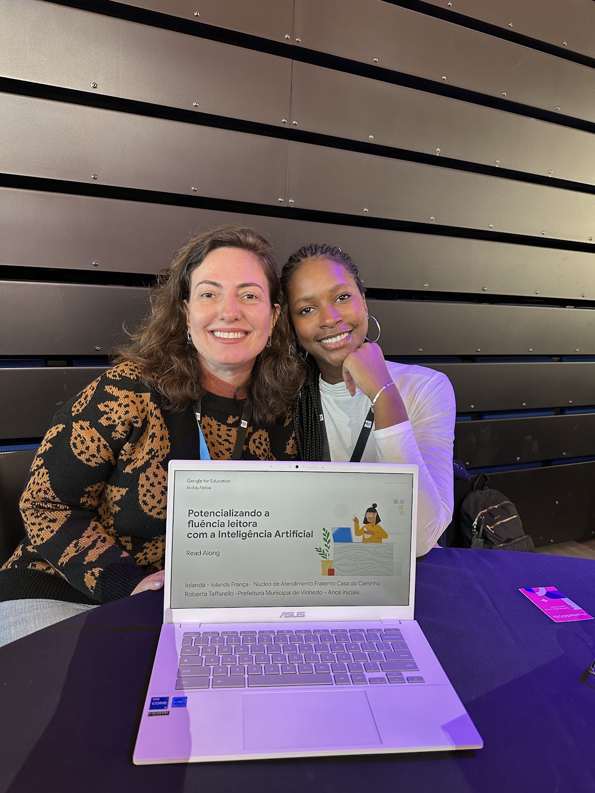 Foto de Roberta Taffarello e Iolanda França sentadas uma ao lado da outra e com um computador na frente delas. Na tela do computador é possível ler o título do trabalho apresentado: Potencializando a fluência leitora com a Inteligência Artificial.