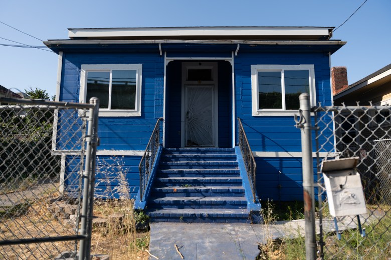 An open wired gate leads to the iconic Warriors House, formerly painted blue and yellow to honor the Golden State Warriors, in Oakland on July 22, 2024. The house was previously a community land trust property. Photo by Florence Middleton, CalMatters