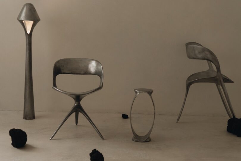four metallic modern furniture pieces—a lamp, a chair, a lounge chair and a side table—are arranged on a neutral floor with dark rocks scattered around, set against a plain beige wall.