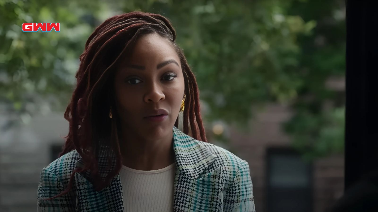 Meagan Good with long dreadlocks wearing a plaid blazer, looking thoughtful.