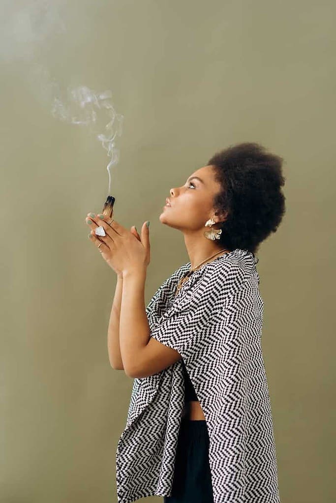 A person is holding a bundle of smoking herbs or incense, with smoke rising from it. The person is wearing a patterned shawl and earrings. The background is a plain, muted green color. This image appears to depict a ritual or spiritual practice involving the burning of herbs or incense, often used for cleansing, meditation, or ceremonial purposes.