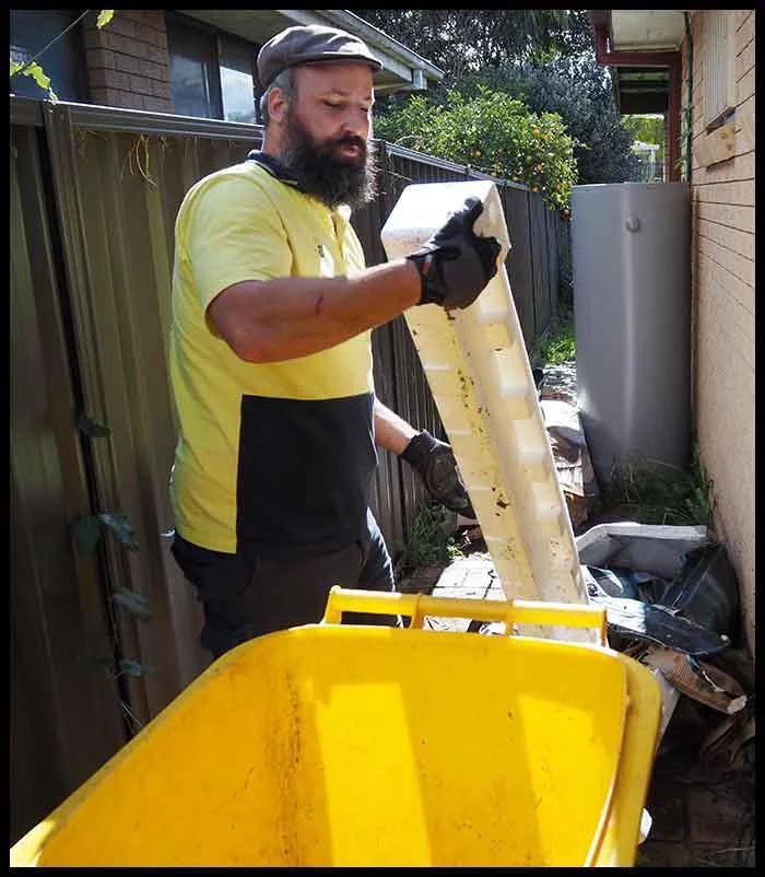 The image shows a Goodbye Junk personnel working outdoors in what appears to be a backyard or side yard area. The individual has a beard and is handling what looks like a panel or board, which they are placing into or removing from a large yellow container that resembles a waste bin or recycling bin.