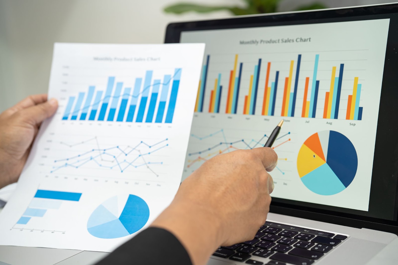 Person holding a pen and a paper with data and looking at a laptop screen with graphs and charts. 