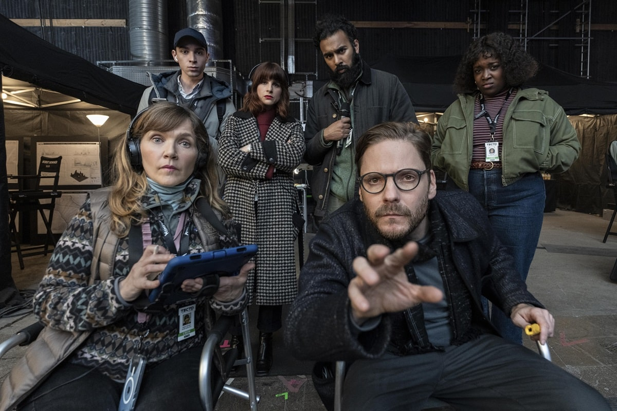 Grupo de personajes de La Franquicia en un set de rodaje. En el centro, Daniel Brühl, sentado, extiende la mano hacia la cámara mientras Jessica Hynes, a su izquierda, sostiene una tableta. A su alrededor, varios personajes más observan con seriedad, todos vestidos con atuendos casuales y etiquetas de identificación del equipo de producción.