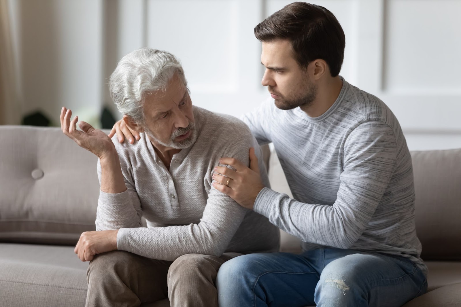 An adult son comforting his parent with his hands on his shoulder and arm when listening to his concerns while sitting on a couch.
