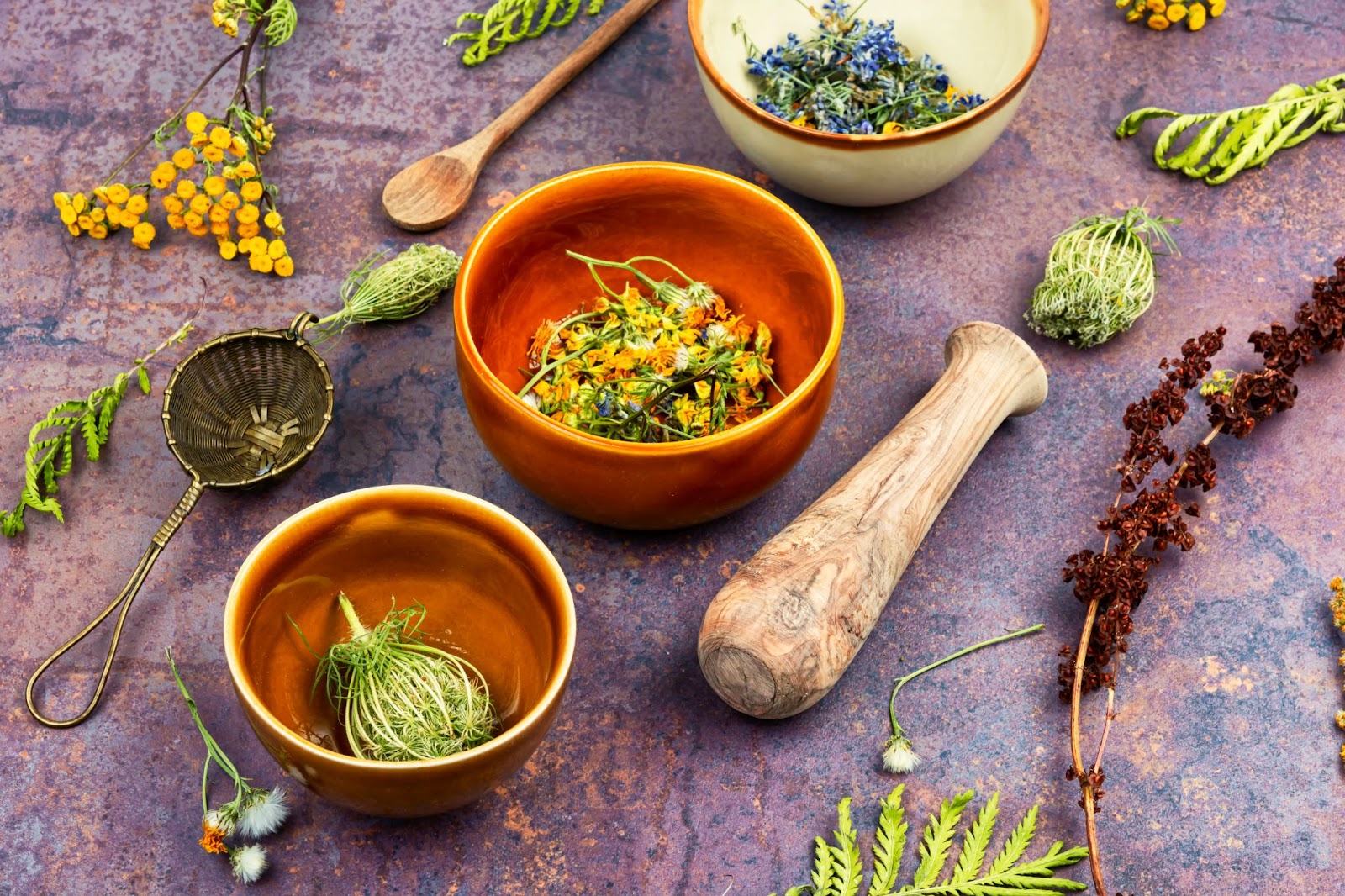 Different herbs in mixing bowls and some scattered on the table alongside a wooden spoon, a strainer, and a pestle