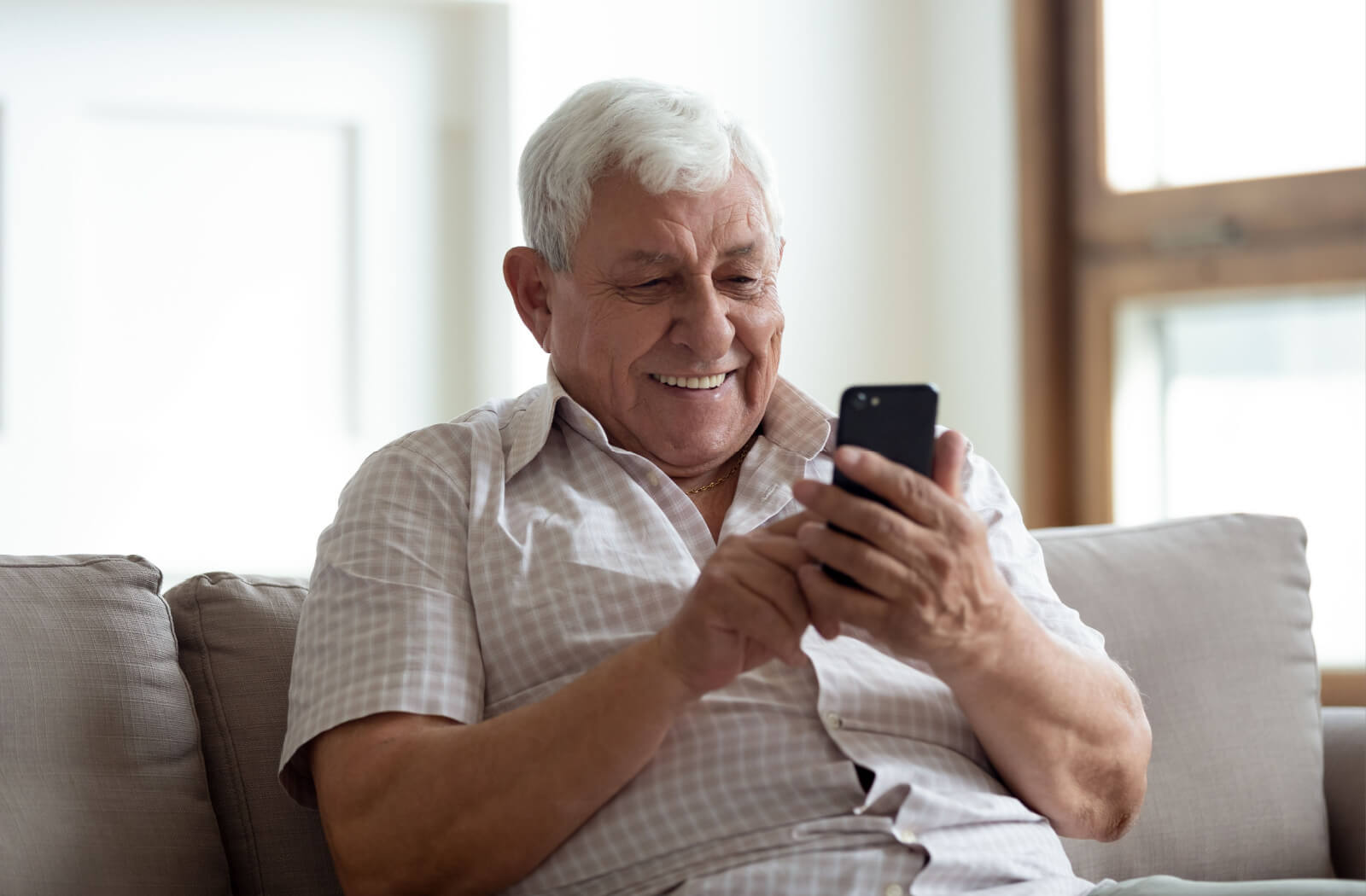 An older adult sitting on their couch, holding their phone and smiling while looking through their apps.