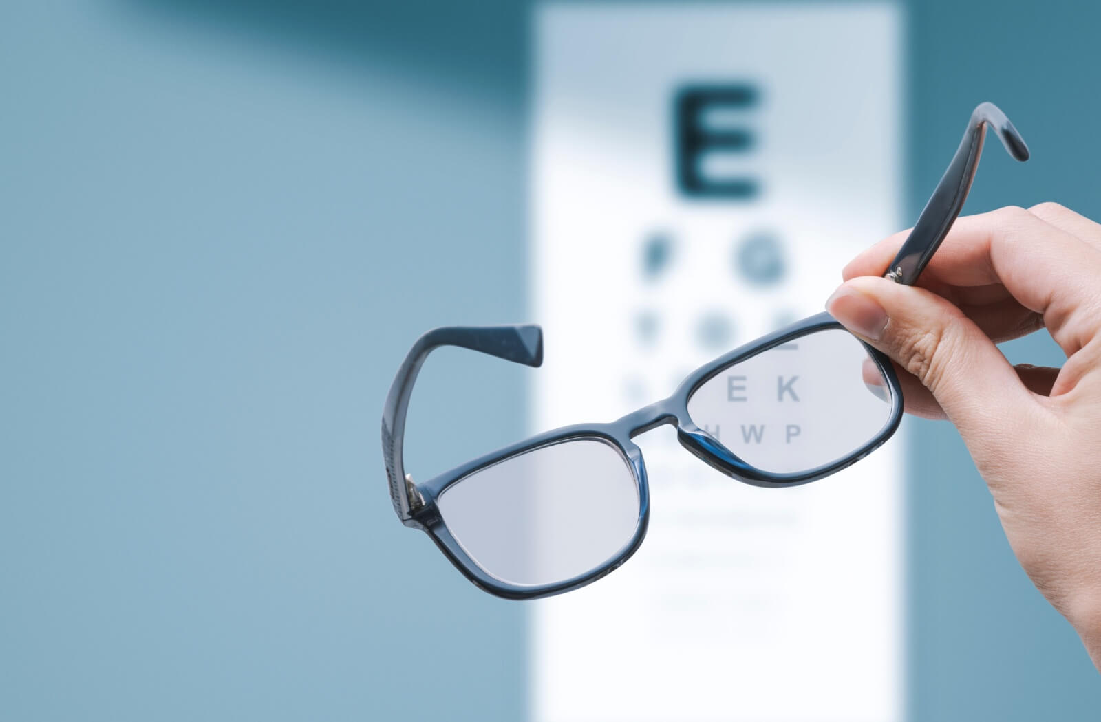 A person holds eyeglasses up to a vision chart. The chart is blurry but the letters seen through the glasses lens are clear.