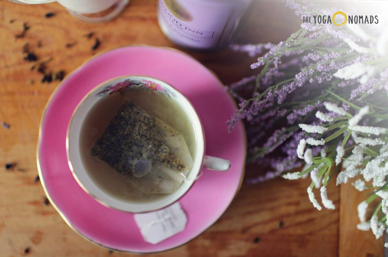 A close-up view of a cup of tea with a tea bag inside, placed on a saucer. To the right side, there is a blurred bunch of flowers, adding an aesthetic and calming vibe. The cup and saucer are on a wooden surface, which appears to be a table. In the background, there is an indistinct object that could be another cup or container.