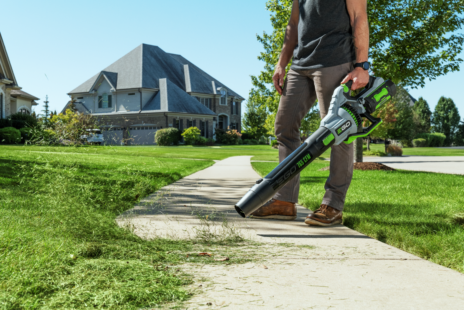 A homeowner easily clearing grass clippings from a sidewalk using an EGO cordless leaf blower in a tidy residential neighborhood, demonstrating quick yard cleanup.