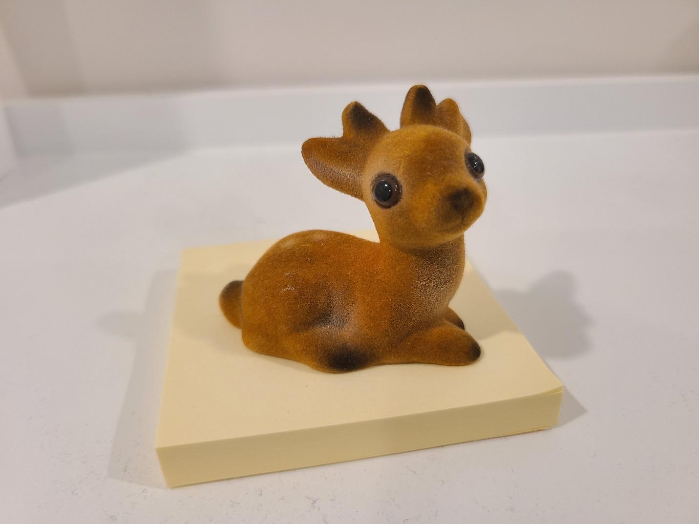 A tiny fuzzy figurine of a brown deer sat down, with two beady black eyes encircled by brown iris, a hint of white spots on its back, and an innocent expression. Photographed on a white shelf