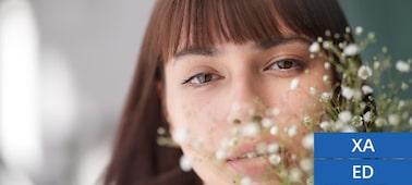 A portrait photograph of a white woman with bangs and flowing hair, facing the camera directly, with a beautiful bokeh effect created by the presence of baby's breath flowers in front of her face.