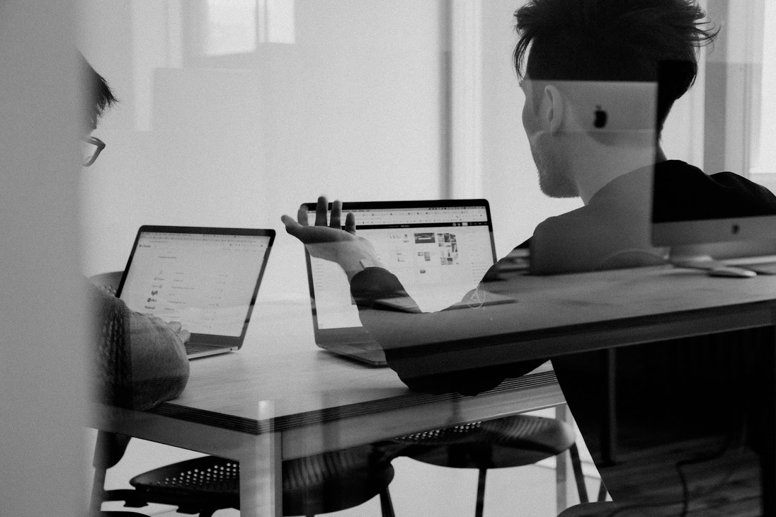 This black-and-white image shows two people sitting at a desk, each working on multiple laptop screens.