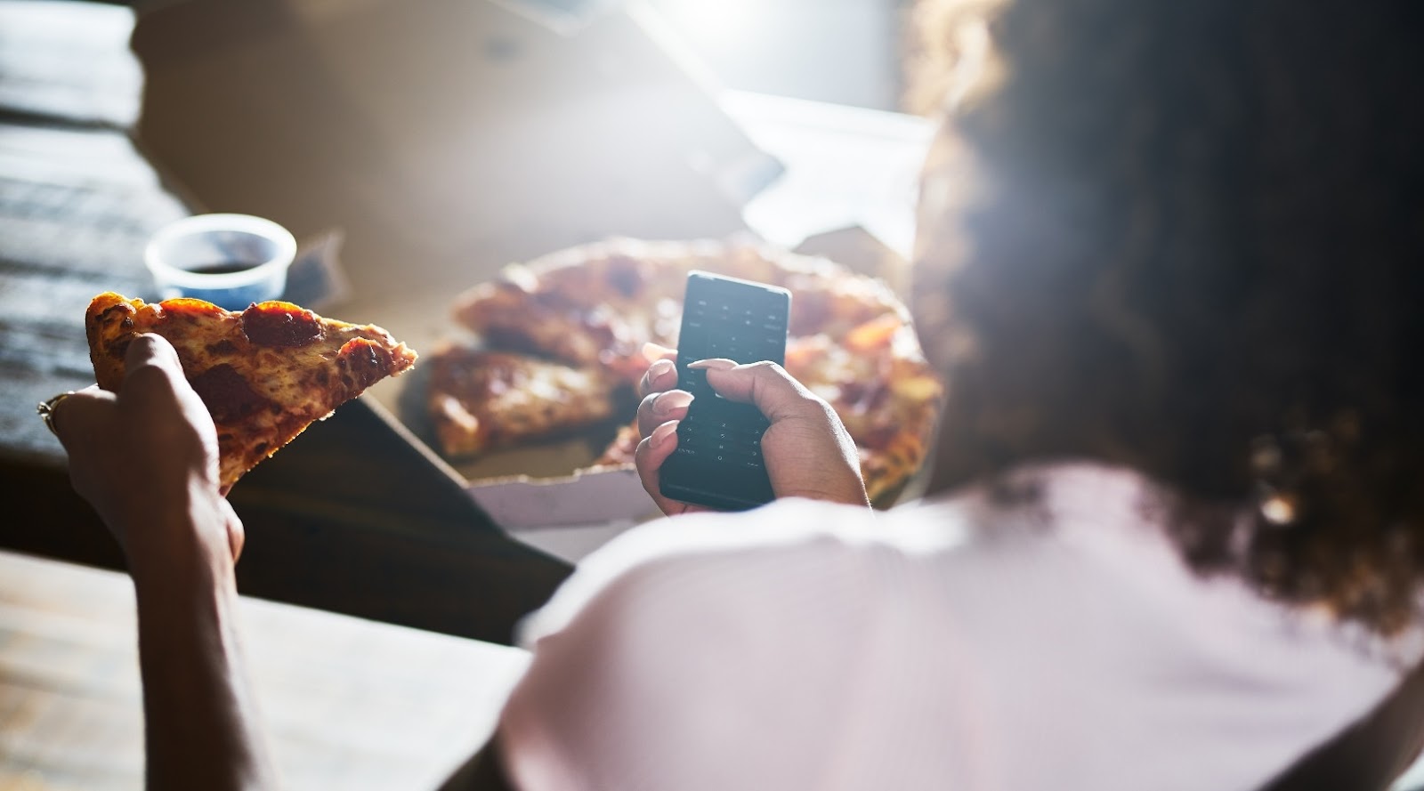 Personne tenant une part de pizza au pepperoni tout en utilisant une télécommande, avec une boîte à pizza ouverte en arrière-plan.