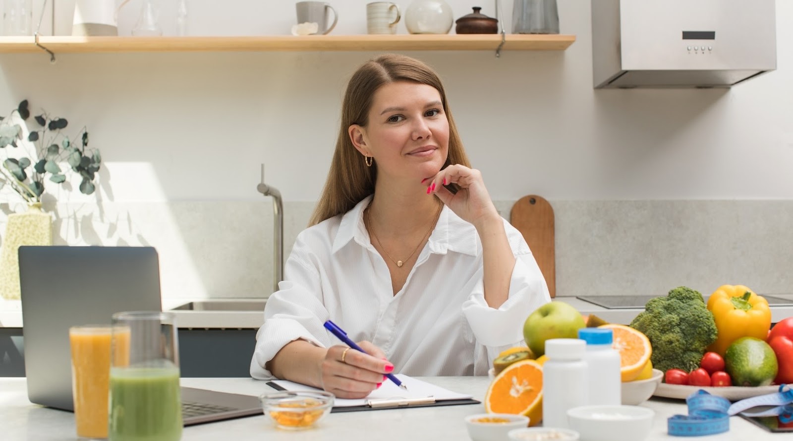 Une diététiste-nutritionniste prend des notes au comptoir de cuisine, entourée de fruits, de légumes frais, de suppléments et d'un ordinateur portable.