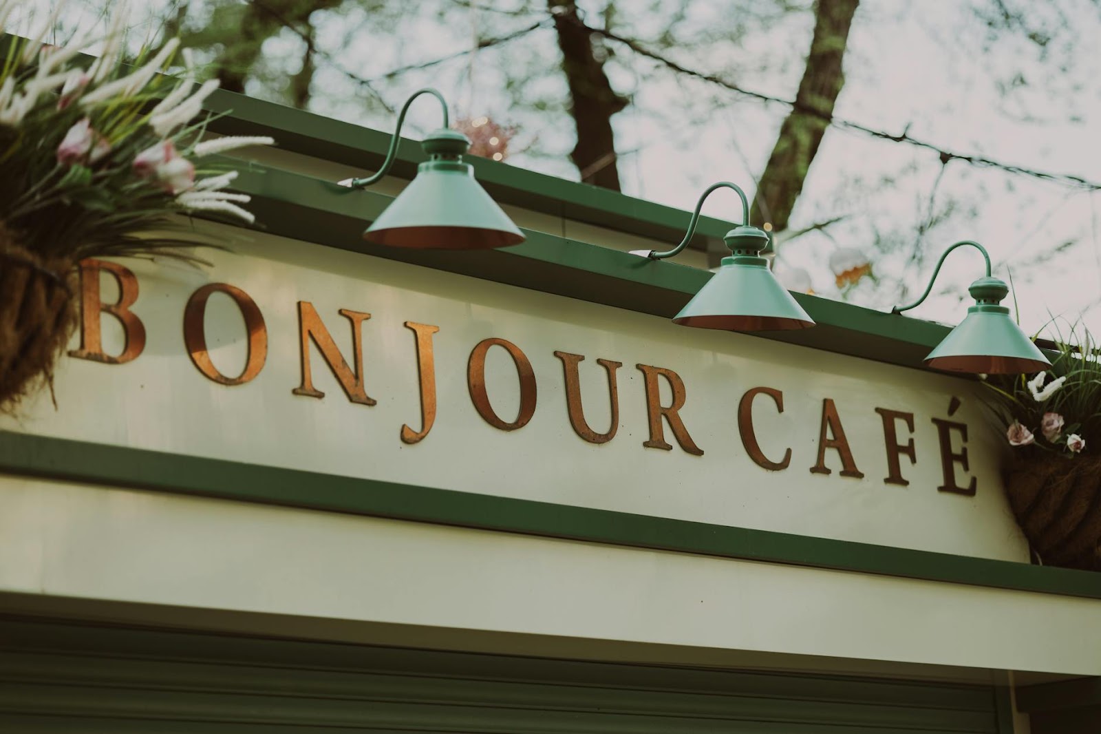 Уличный снимок вывески Bonjour Café со старинными лампами и цветами.