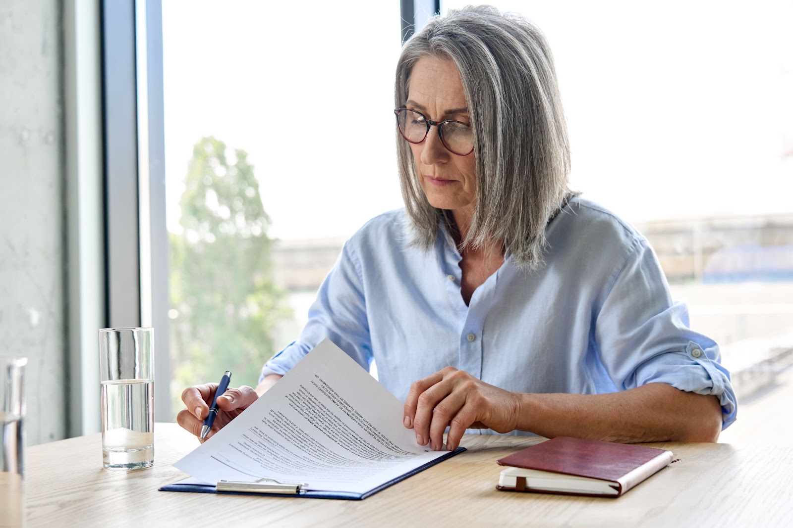 Older woman wearing eyeglasses signs power of attorney document at desk in an office.