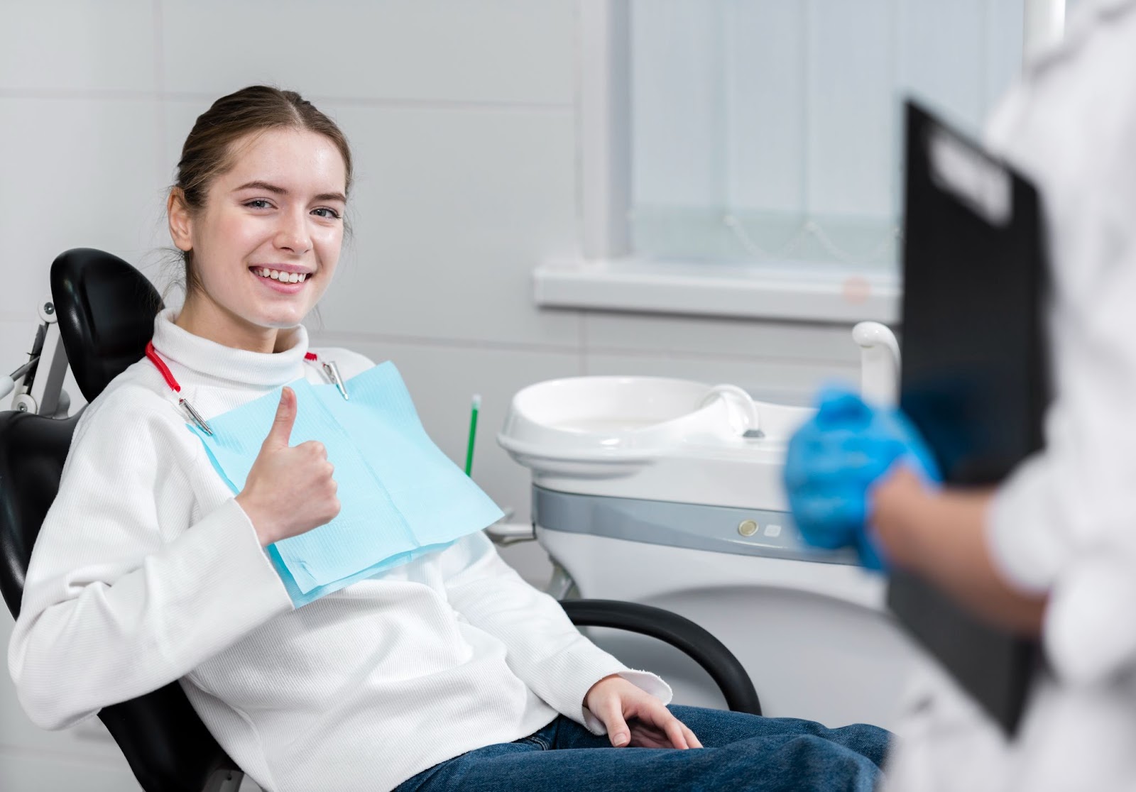 Smiling young woman giving a thumbs up while sitting in a dental chair after treatment, wearing a protective bib, showing satisfaction with dental care.