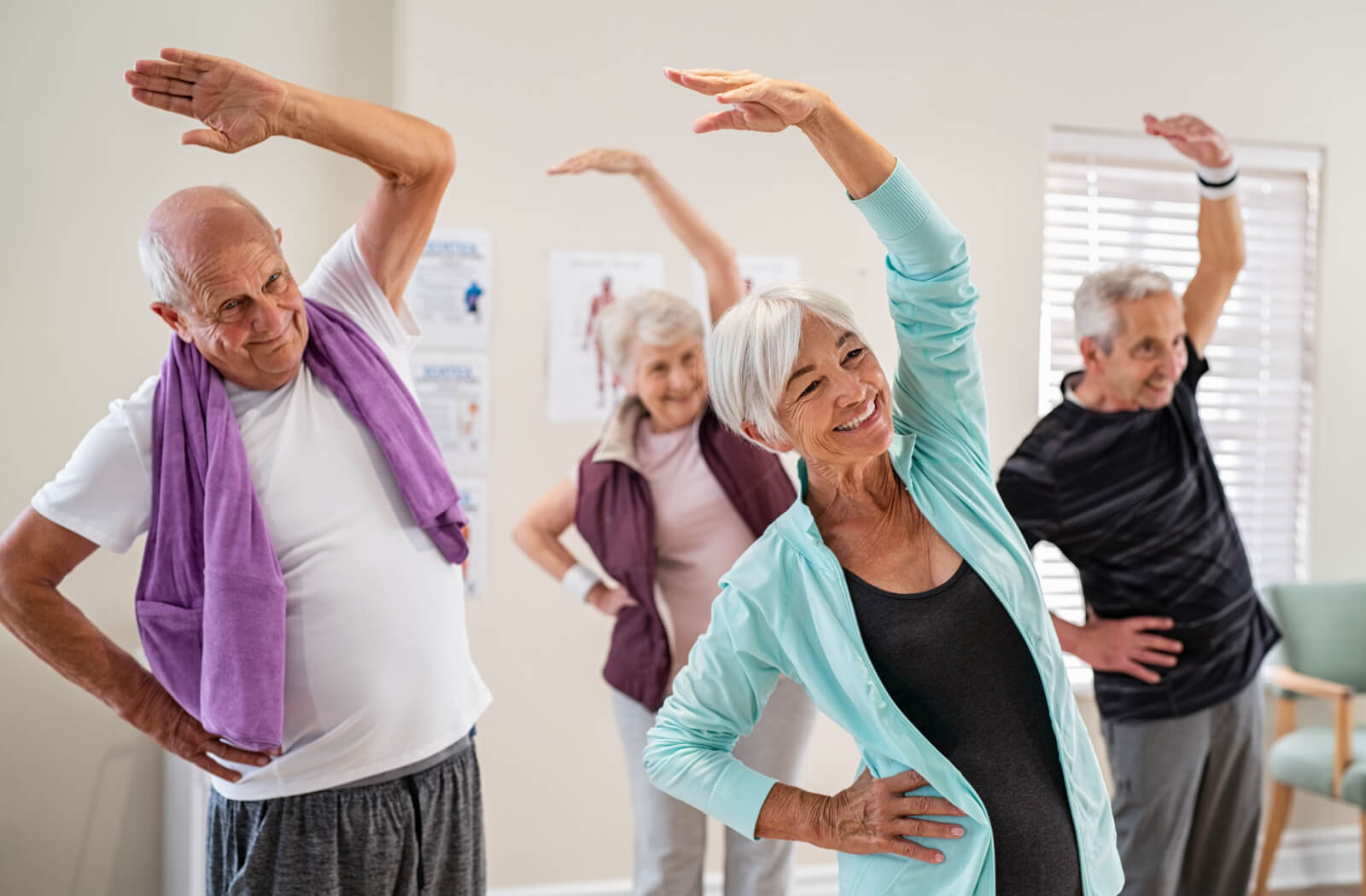 4 seniors indoors in a fitness center smiling and stretching their arms overhead during a physical exercise class.