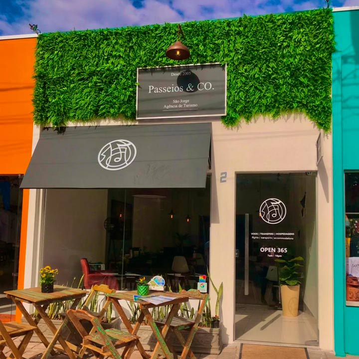 Imagem de uma loja chamada Passeios & Co., com fachada decorada com verdura artificial e móveis de madeira na frente.
