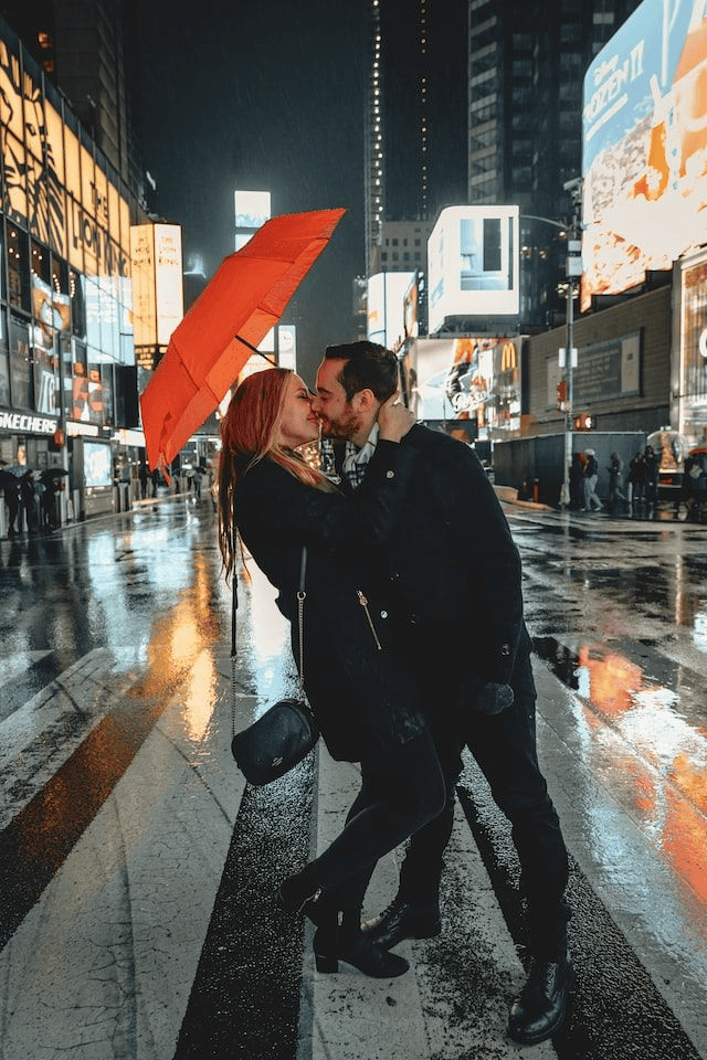 Rainy photoshoot featuring a couple passing a kiss under umbrella, smiling and radiating happiness