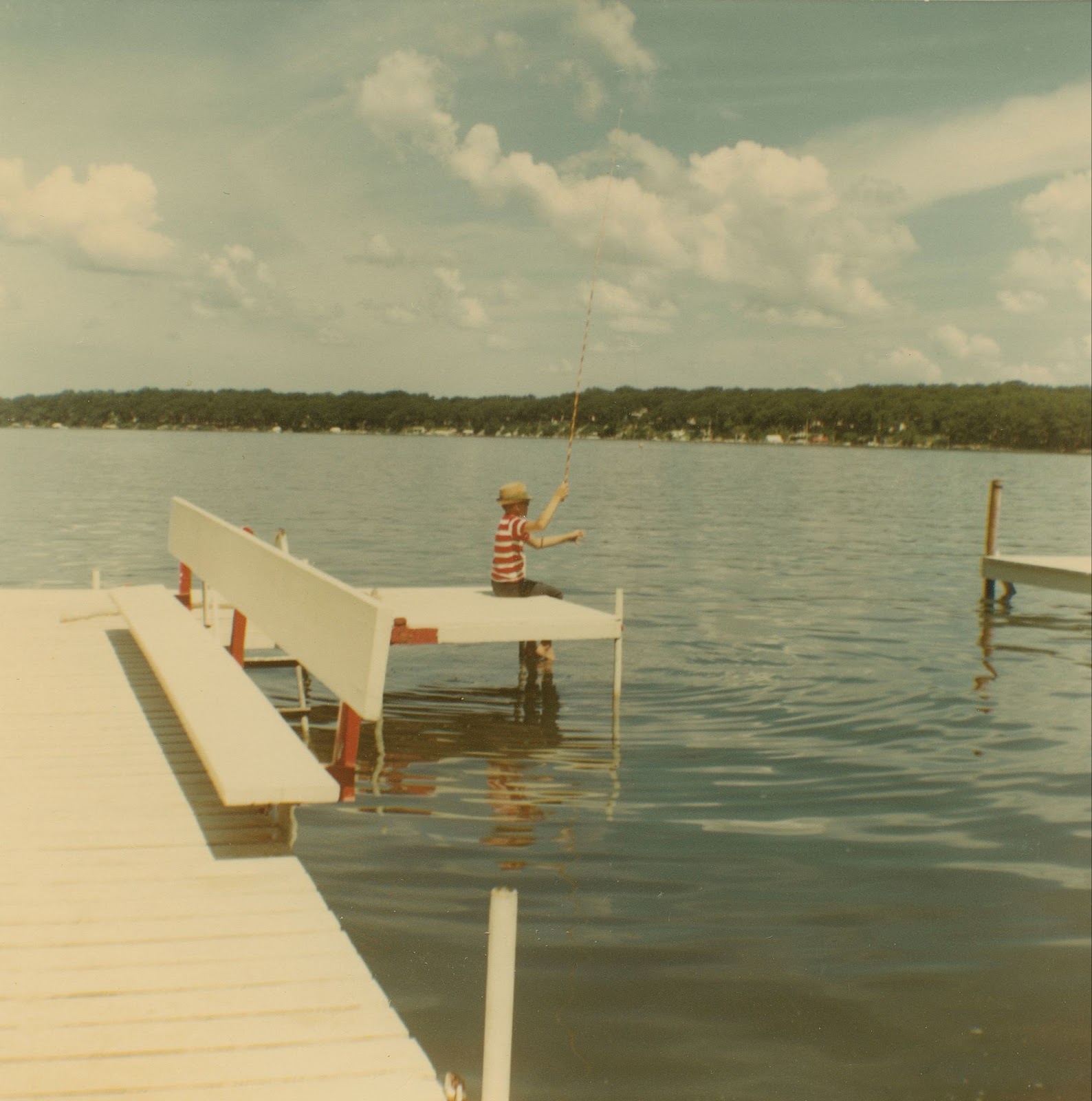 Little boy wearing a red and white striped shirt sits at the end of a dock, fishing. 