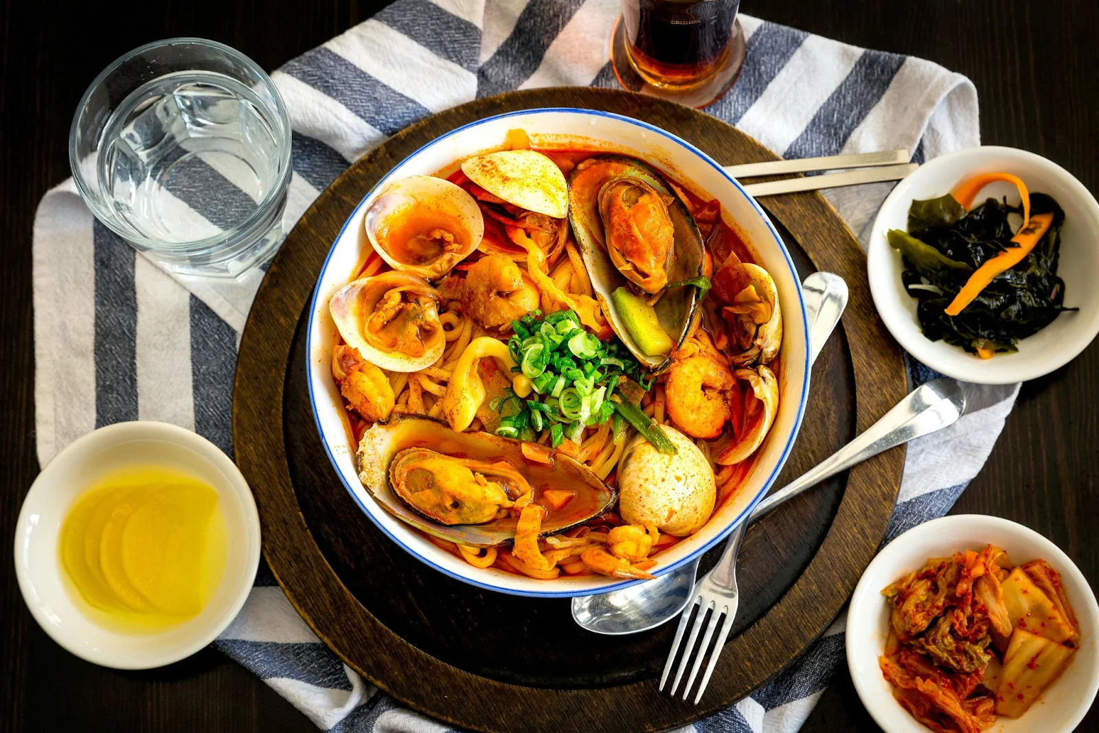 korean kimchi in bowl with cutlery. sauces and oil is also placed along. fork and spoon and glass of water.