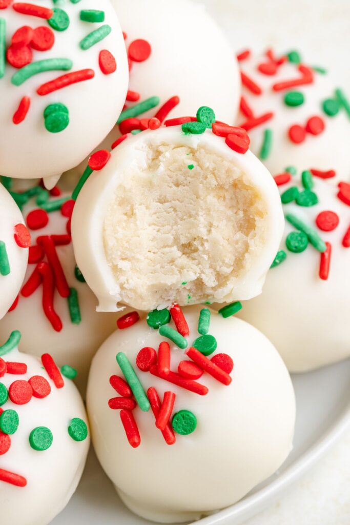 Truffles with sugar cookies, topped with red and green splashes, are stacked on a plate. One trumel is cut, showing the cream interior.