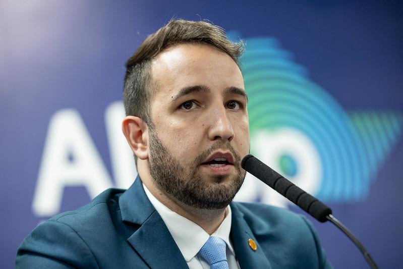 Foto de homem branco com barba e cabelos castanhos falando ao microfone. Ele usa terno e gravata. O fundo é azul. 