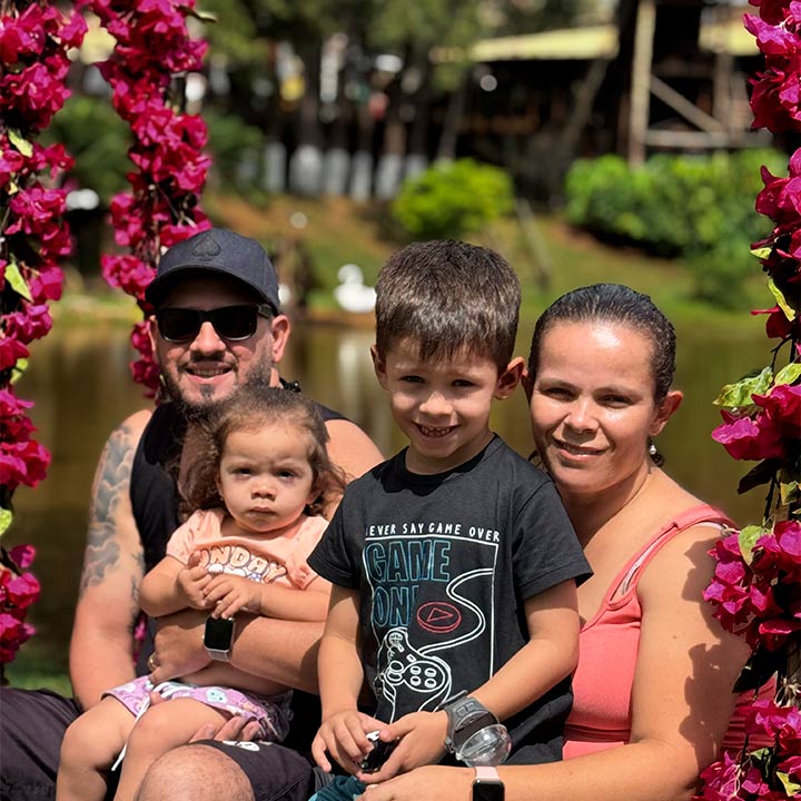 Família sorridente em um passeio ao ar livre, cercada por flores rosadas, com crianças e adultos felizes em um dia ensolarado na natureza.