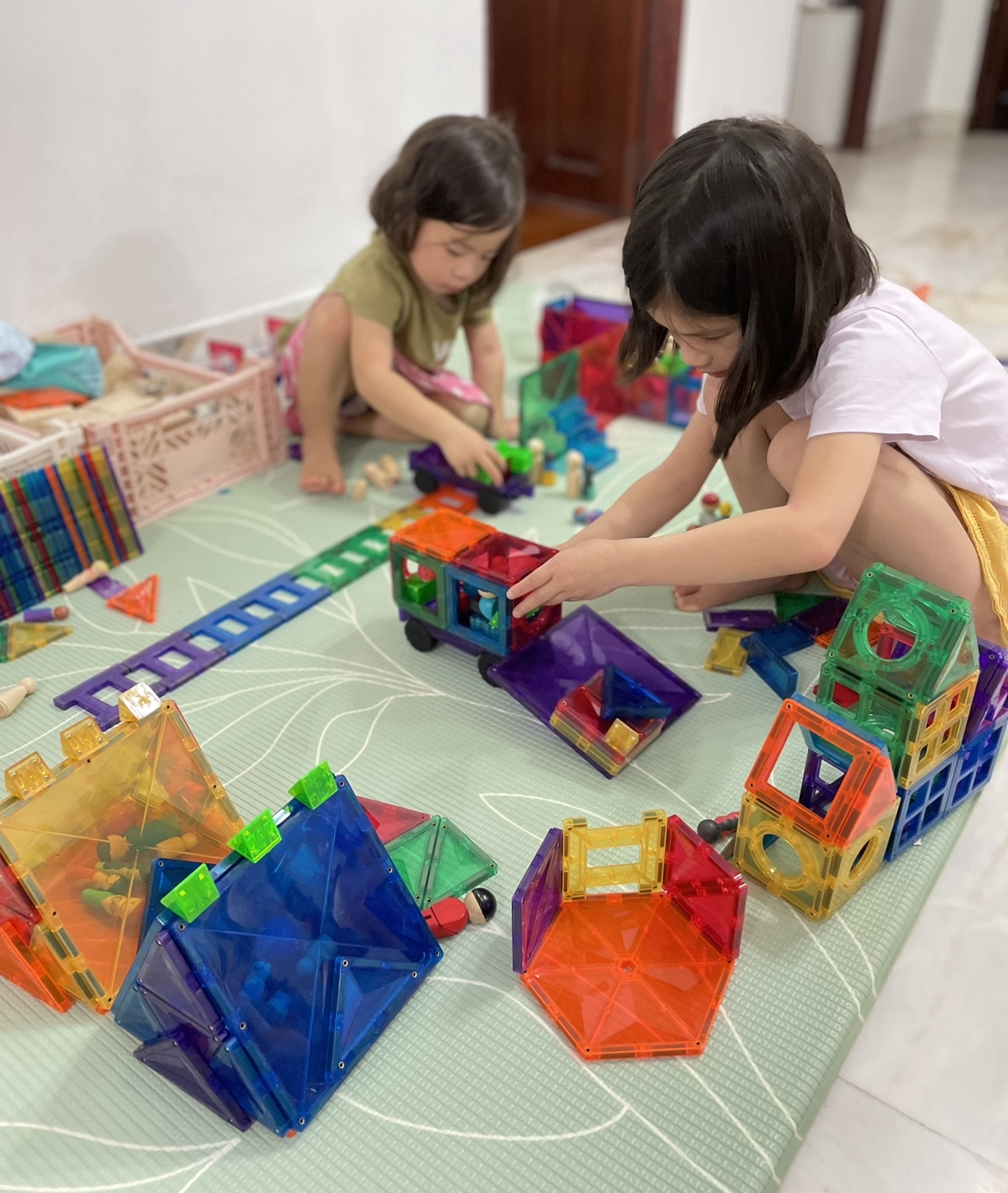 Siblings, a toddler and preschooler, playing side by side, each immersed in their own imaginative creations—one building a dump truck and the other playing with a car on a track. Together, they use colorful magnetic tiles to build a city with structures and roads, showcasing the open-endedness of magnetic tiles that fosters creativity, teamwork, and limitless possibilities for exploration.