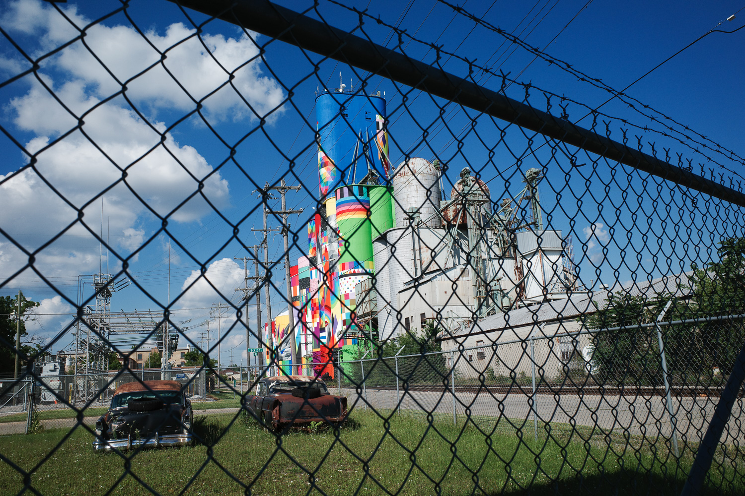 Distant view of Shine Bright Saginaw mural with barbed wire fence and rusted cars in foreground.
