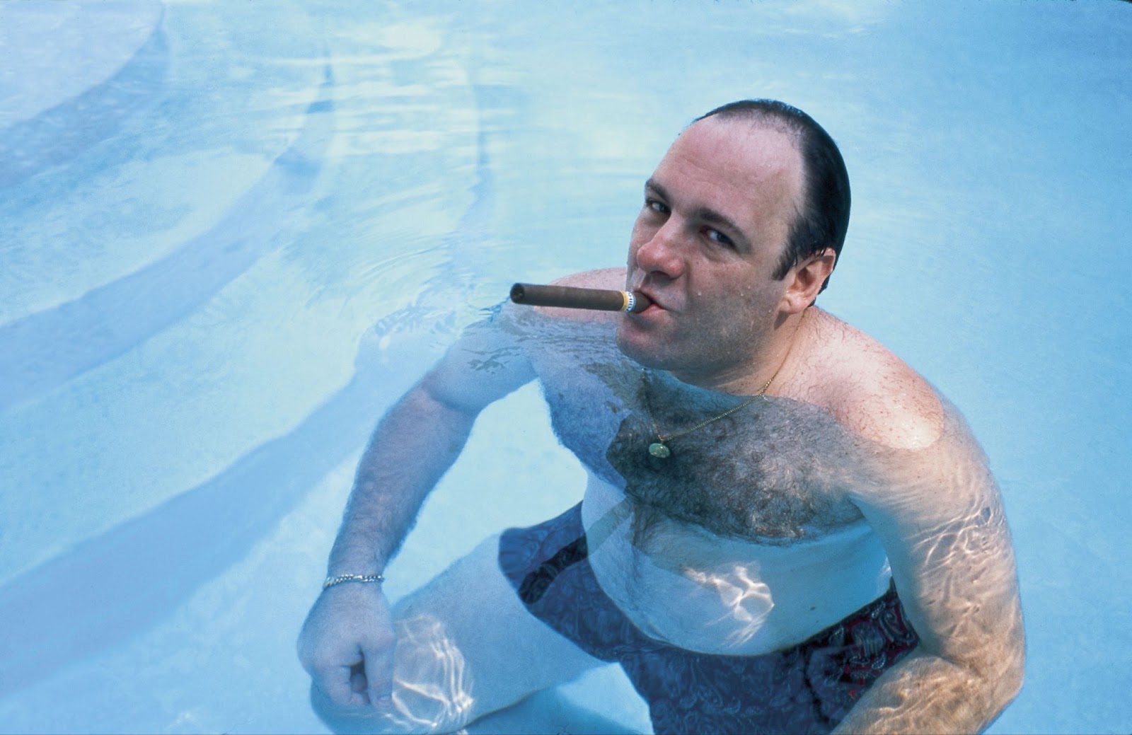 James Gandolfini en su papel icónico de Tony Soprano, relajado en una piscina y fumando un cigarro. Lleva un medallón dorado y trajes de baño, con una expresión relajada y ligeramente desafiante, capturando la esencia de su personaje.