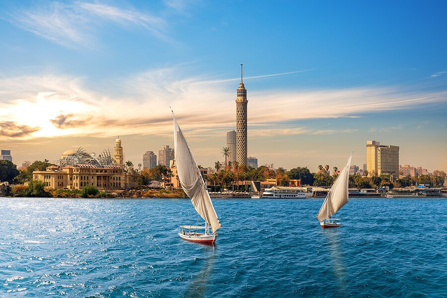 Cairo skyline with the Nile River, traditional sailboats (feluccas), and Cairo Tower under a vibrant blue sky during sunset.