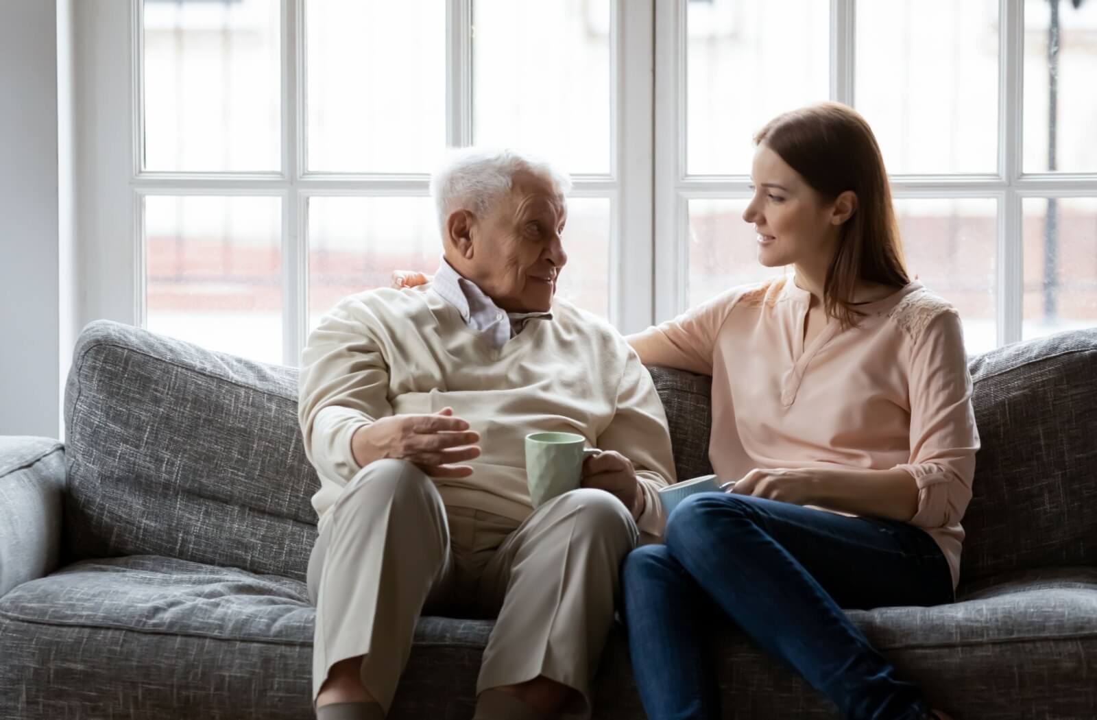 an adult child comforts their senior parent on a grey sofa.
