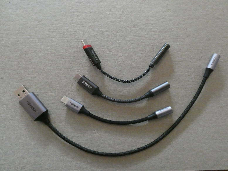 Four USB to 3.5mm dongles with braided cables, one above the other. From top to bottom, USB-C black with a red trim on the USB plug (Cubliux), USB-C black with a silver 3.5mm barrel (Anker), USB-C silver (UGreen) and USB-A silver with a longer cord (Ugreen).