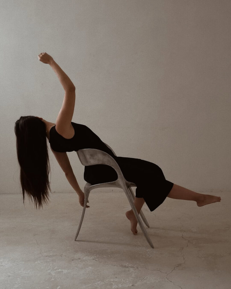 A person in a black outfit leans backward over a metal chair with one arm raised and feet off the ground in a minimalist room