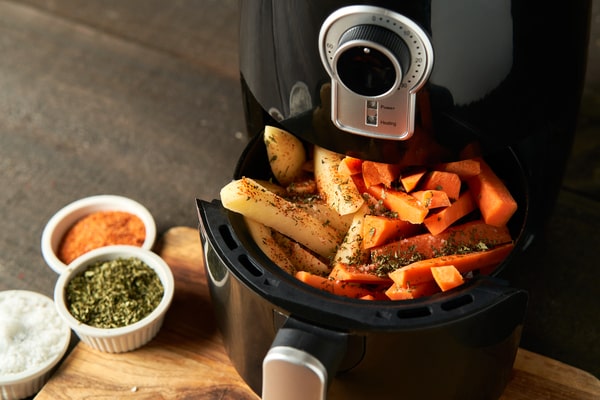 An air fryer basket filled with seasoned sweet potato and white potato wedges, ready to be cooked, with small bowls of spices and herbs on the side